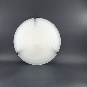 Pode incluir: Uma luminária de teto circular branca com um abajur de vidro fosco e anéis concêntricos. A luminária tem quatro suportes de montagem de metal preto.