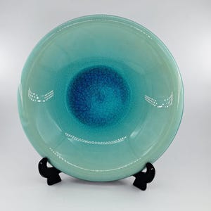 Könnte beinhalten: Eine runde Keramikschale mit einem Farbverlauf, der von einer tiefblauen Mitte mit feiner Rissstruktur zu einem helleren, glatten türkisfarbenen Rand übergeht. Die Schale wird von einem kleinen, schwarzen, zweizinkigen Ständer gehalten.