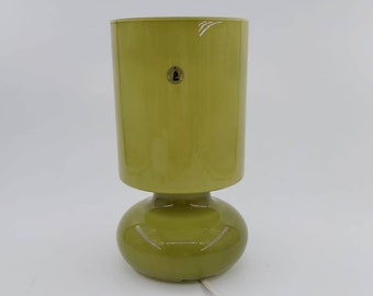 May include: A green table lamp with a cylindrical shade and a rounded base. The lamp has a glossy finish and a white power cord. The lamp is approximately 12 inches tall.