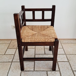 Op de afbeelding: Een donkerbruine houten hoekstoel met een gevlochten zitting van biezen. De stoel heeft een vierkant frame met een rugleuning bestaande uit twee loodrechte delen, elk met drie verticale spijlen, en vier stevige poten met dwarsbalken.