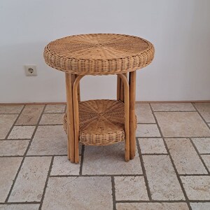Op de afbeelding: Een naturelkleurige, tweelaagse bijzettafel van riet. De tafel heeft een rond blad en een onderste plank, beide geweven met een strak patroon. De poten zijn gemaakt van verticale rieten steunen. De tafel is ongeveer 61 cm hoog.