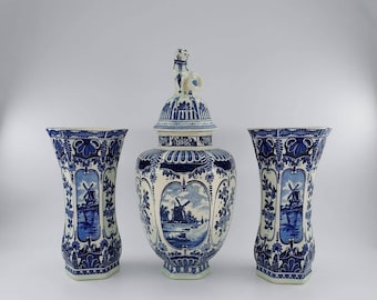 May include: A set of three blue and white Delftware vases. The largest vase has a lid with a small white dog figurine on top. All three vases feature a windmill scene with blue and white floral designs.