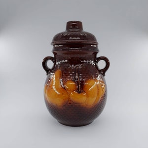 May include: A brown ceramic jar with a lid, decorated with a raised design of pears and cherries. The jar has two handles on the sides.
