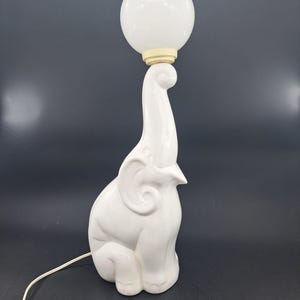 Op de afbeelding: Een witte keramische olifant tafellamp met een glanzende afwerking en een gestileerde slurf die omhoog wijst. De lamp is voorzien van een bolvormige witte glazen lamp die in een crèmekleurige fitting rust. Een wit snoer komt uit de basis.