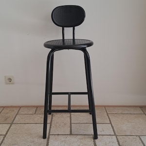 May include: A black metal bar stool with a round seat and a back rest. The stool is standing on a tiled floor.