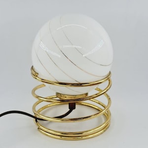 Op de afbeelding: Een witte bollamp met gouden strepen staat op een gouden metalen spiraalvormige voet. De lamp heeft een snoer.