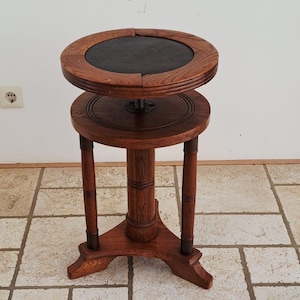 Pode incluir: Um suporte de pedestal de madeira castanha com um topo circular e um centro escuro embutido. O suporte tem uma coluna central apoiada por três pernas e inclui um componente de ajuste mecânico entre o topo e a estrutura da base.