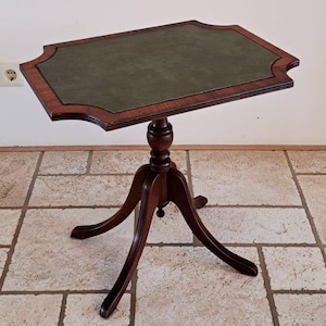 Op de afbeelding: Een kleine, antieke bijzettafel met een donkere houten afwerking. Het tafelblad is donkergroen van kleur, omlijst door een decoratieve rand. De tafel heeft een voetstuk met drie gebogen poten. De tafel is ongeveer 71 cm hoog.