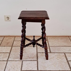 May include: A dark brown wooden stool with a square top and four legs featuring a twisted barley sugar design. The legs are connected by a central cross-shaped stretcher with a small decorative finial at the intersection point.