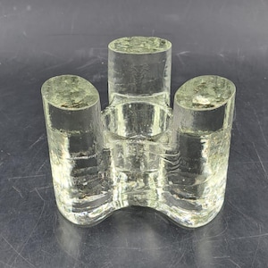 Puede incluir: Un portavelas de vidrio transparente con tres aberturas cilíndricas. El portavelas tiene una forma única y orgánica y está diseñado para sostener tres velas.