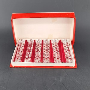 Op de afbeelding: Een set rode en heldere glazen kandelaars in een rood-witte doos. De kandelaars hebben een decoratief patroon en zijn netjes in de doos gerangschikt. De doos is open en onthult de inhoud.
