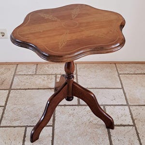 May include: A small, wooden side table with a unique, scalloped tabletop. The table is brown with a decorative inlaid design on the top. It has a pedestal base with three curved legs. The table is approximately 61 cm tall.