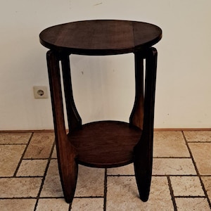 Puede incluir: Una mesa auxiliar de madera de color marrón oscuro con una superficie circular y un estante inferior circular más pequeño. La mesa se apoya en tres patas verticales curvas que se extienden desde la parte superior hasta la base.