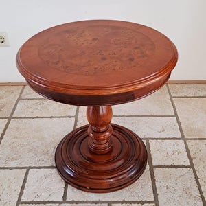 Op de afbeelding: Een ronde houten bijzettafel met een donkerbruine afwerking. De tafel heeft een cirkelvormig blad met een decoratief houtnerfpatroon en een voetstuk. De tafel is ongeveer 61 cm in diameter en 71 cm hoog.