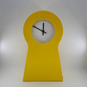 May include: A bright yellow table clock with a keyhole-shaped frame. The clock face is circular and white, featuring thin black hour and minute hands, along with small black numerical markings for each hour positioned around the perimeter.