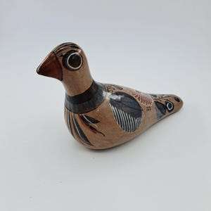 Op de afbeelding: Een handbeschilderd keramieken vogelbeeldje in een lichtbruine kleikleur. De vogel heeft een donkerbruine snavel, een zwarte nekband en ingewikkelde zwart-witte lijnpatronen op de vleugels en het lichaam, geaccentueerd met een kleine rood-oranje bloem.
