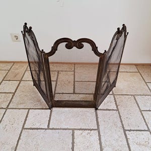Può includere: Un parascintille a tre pannelli in metallo scuro con una rete a maglia fine. Il pannello centrale presenta una parte superiore ad arco decorativa con dettagli a voluta, mentre i due pannelli laterali sono incernierati per il posizionamento.