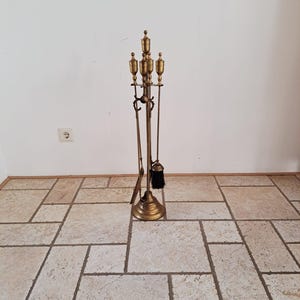 Könnte beinhalten: Ein Kamingarnitur-Set aus Messing mit einem zentralen Ständer auf einem runden Fuß, das eine Schaufel, einen Schürhaken und eine Bürste mit schwarzen Borsten hält. Jeder Werkzeuggriff ist mit einem dekorativen Endstück aus Messing versehen.
