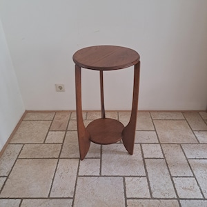 Pode incluir: Um suporte de plantas de madeira com um tampo redondo e uma prateleira inferior. O suporte tem três pernas curvas e é feito de madeira de tom médio. O suporte tem aproximadamente 71 cm de altura.