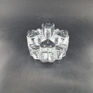 Puede incluir: Un portavelas de vidrio transparente con forma de trébol de cuatro hojas con un centro elevado para una vela. El portavelas tiene una etiqueta "Dacono" en la parte superior.