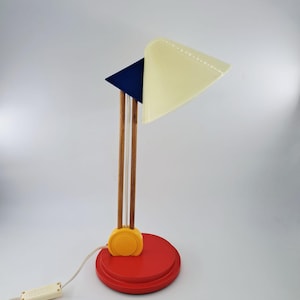 Op de afbeelding: Een tafellamp met een rode ronde voet, een geel scharnier, twee verticale houten stelen, een donkerblauw driehoekig accentstuk en een crèmekleurige conische kap. Een wit snoer met een schakelaar loopt vanaf de voet naar buiten.