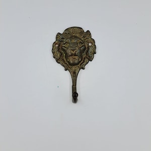 Op de afbeelding: Een wandhaak van gegoten metaal met een gedetailleerd leeuwenkopontwerp in een donkere, verweerde bronsafwerking. Het leeuwengezicht dient als decoratieve achterplaat, met een gebogen haak die naar beneden uitsteekt om voorwerpen op te hangen.