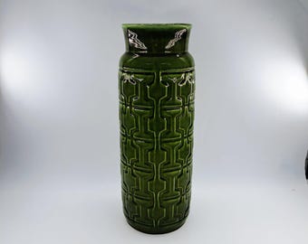 Può includere: Un vaso alto in ceramica verde con un motivo geometrico. Il vaso ha una forma cilindrica con una parte superiore leggermente svasata e una finitura lucida. Il motivo è un disegno ripetuto di forme interconnesse. Il vaso è alto circa 30 cm.