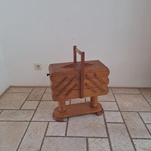 May include: A vintage wooden sewing box with a handle, featuring multiple compartments that open outwards. The box is made of light-colored wood and stands on a decorative base. The box is designed for storing sewing supplies.