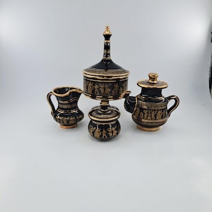 Op de afbeelding: Een zwart en goud keramisch theeservies met een melkkan, theepot, suikerpot met deksel en een pot met deksel. De set heeft gouden accenten en Grieks geïnspireerde figuren en patronen. De theepot heeft een handvat en een tuit.