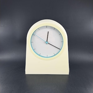 Op de afbeelding: Een witte analoge klok met een blauwe en gele rand rond de wijzerplaat. De klok heeft zwarte wijzers en staat op een zwart oppervlak.