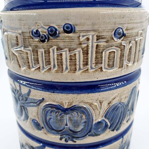 Peut inclure: Un pot en c&eacute;ramique blanche avec des accents bleus et bruns. Le pot a un motif d&eacute;coratif de fruits et l&eacute;gumes, et le mot "Kuntopf" est &eacute;crit sur le c&ocirc;t&eacute;.
