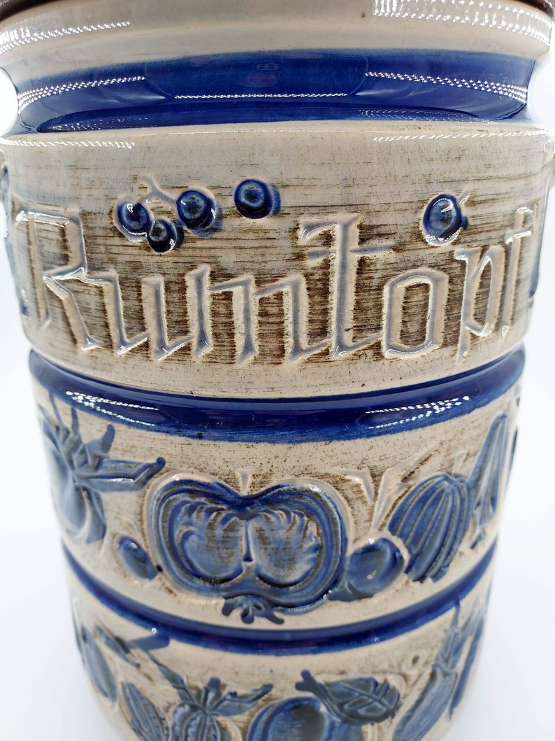Peut inclure: Un pot en c&eacute;ramique blanche avec des accents bleus et bruns. Le pot a un motif d&eacute;coratif de fruits et l&eacute;gumes, et le mot "Kuntopf" est &eacute;crit sur le c&ocirc;t&eacute;.