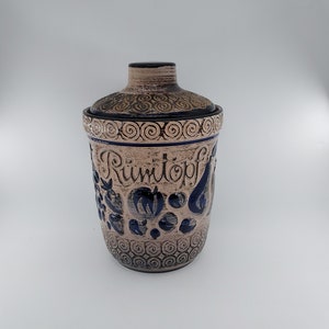 May include: A ceramic jar with a lid, decorated with blue and gold swirls and floral designs. The jar is labeled "Rumtopf" in German.