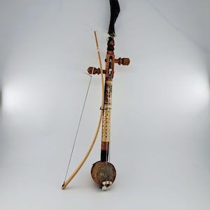 Può includere: Uno strumento musicale a corde con una base risonante rotonda rivestita in pelle, un lungo manico in legno e un terminale superiore curvo. Presenta un archetto in legno chiaro con una sottile corda nera, piroli e intarsi decorativi.