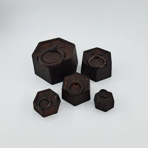 Puede incluir: Un conjunto de cinco pesas de metal de color marrón oscuro, con forma hexagonal y tamaños variados, cada una con un asa circular en la parte superior. Las pesas están grabadas con sus masas respectivas: 5kg, 2kg, 1kg, 1/2kg y 1/4kg.