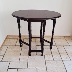May include: A dark brown, round, wooden drop-leaf table. The table has a dark finish, with a circular top and four legs. The legs are intricately carved with a twisted rope design. The table is approximately 76 cm in diameter when fully extended.