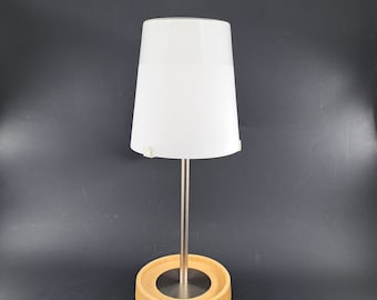 May include: A contemporary table lamp featuring a white lampshade and a brushed metal stem. The lamp is set on a circular, light-coloured wooden base. A white power cord extends from the base.