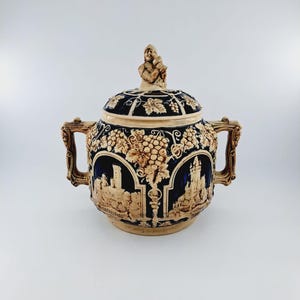 May include: A ceramic lidded jar with two handles, decorated with a blue and white glaze and intricate relief carvings of grapes, vines, and a castle. The lid features a small figurine of a man holding a cup.