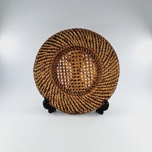 May include: A round woven wicker plate with a decorative woven pattern. The plate is brown and has a natural finish. It is displayed on a black stand.