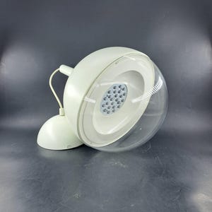 Op de afbeelding: Een witte bureaulamp in retrostijl met een bolvormige kop en een transparante, gewelfde kap. De lamp heeft een cirkelvormige reeks kleine lampjes en een gebogen arm. De basis is rond.