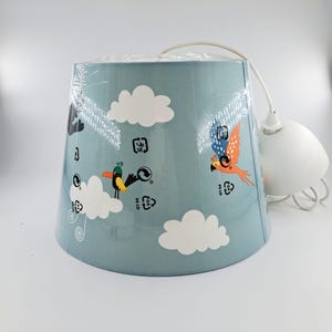 May include: A light blue lampshade with a whimsical design featuring white clouds and colorful birds. The lampshade is cylindrical and has a white cord and fixture attached. The design includes a parrot with orange and blue wings.