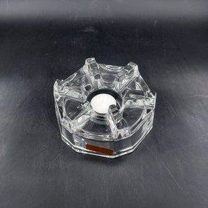 Puede incluir: Un portavelas de cristal transparente y facetado con un diseño geométrico. El portavelas tiene un espacio circular central para una pequeña vela blanca. Una pequeña etiqueta rectangular es visible en el lateral. El portavelas está sobre una superficie oscura.