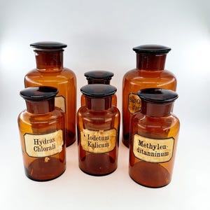 Op de afbeelding: Vijf amberkleurige glazen flessen met zwarte deksels en vintage etiketten. De etiketten hebben tekst in het Duits, waaronder "Hydras Chlorali", "Iodetum Kalicum" en "Methylen ditanninum".