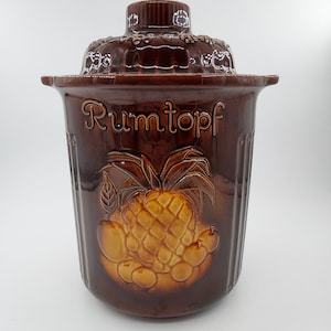 May include: A brown ceramic crock with a lid, featuring a pineapple and other fruits in relief. The crock is labelled "Rumtopf" in gold lettering.