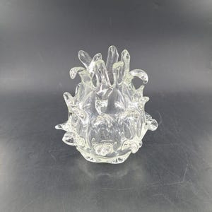 Op de afbeelding: Een decoratieve kom van helder glas met een organisch, stekelig ontwerp. De kom heeft een afgeronde basis en talrijke opwaarts gerichte, bloembladachtige uitsteeksels. Het glas is transparant en staat op een donkere ondergrond.