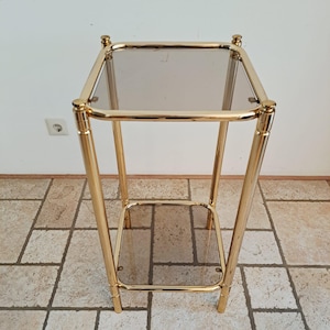 Puede incluir: Una mesa auxiliar cuadrada con un marco de metal en tono dorado pulido y dos estantes de cristal ahumado. La mesa cuenta con cuatro patas verticales con remates redondeados en las esquinas superiores y un estante inferior a juego.
