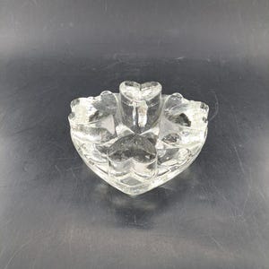 Puede incluir: Un portavelas transparente en forma de corazón con cinco compartimentos en forma de corazón. El soporte es de vidrio grueso y se asienta sobre una superficie oscura. El diseño es simple y elegante, adecuado para un ambiente romántico.