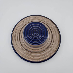 Peut inclure: Une assiette en c&eacute;ramique avec des cercles concentriques dans des tons beige et bleu. L'assiette a un centre sur&eacute;lev&eacute; avec un motif de cercle bleu.