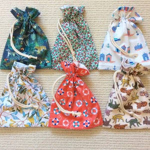 May include: Six fabric drawstring pouches featuring various patterns including a dark jungle print with toucans, a small red floral print, a white beach hut and anchor print, a blue and yellow leaf print, a red lifebuoy print, and a cat print.