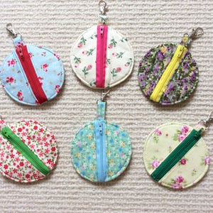 May include: Six circular fabric coin pouches with metal lobster claw clasps. Each pouch features a central zip in a contrasting colour and a floral pattern. The designs include light blue, cream, purple, white, teal, and pale yellow fabric bases.